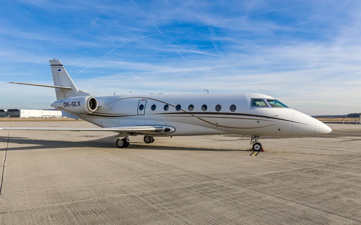 A white Gulfstream G200 business jet with a brown stripe is parked on an airfield under a blue sky.