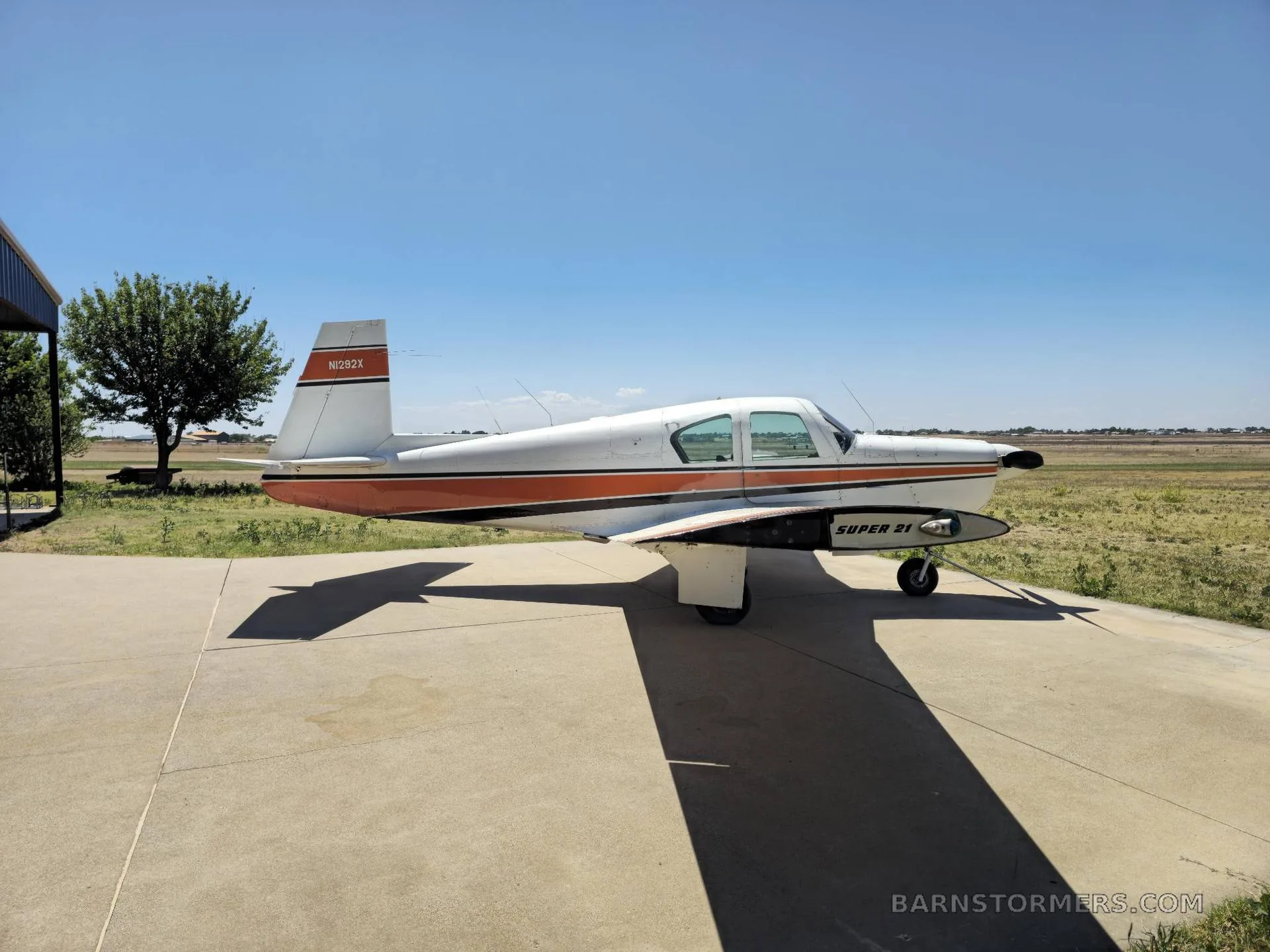 A white and orange single-engine aircraft (N282X) with "SUPER 21" text on its side, parked on a concrete pad under a clear blue sky.