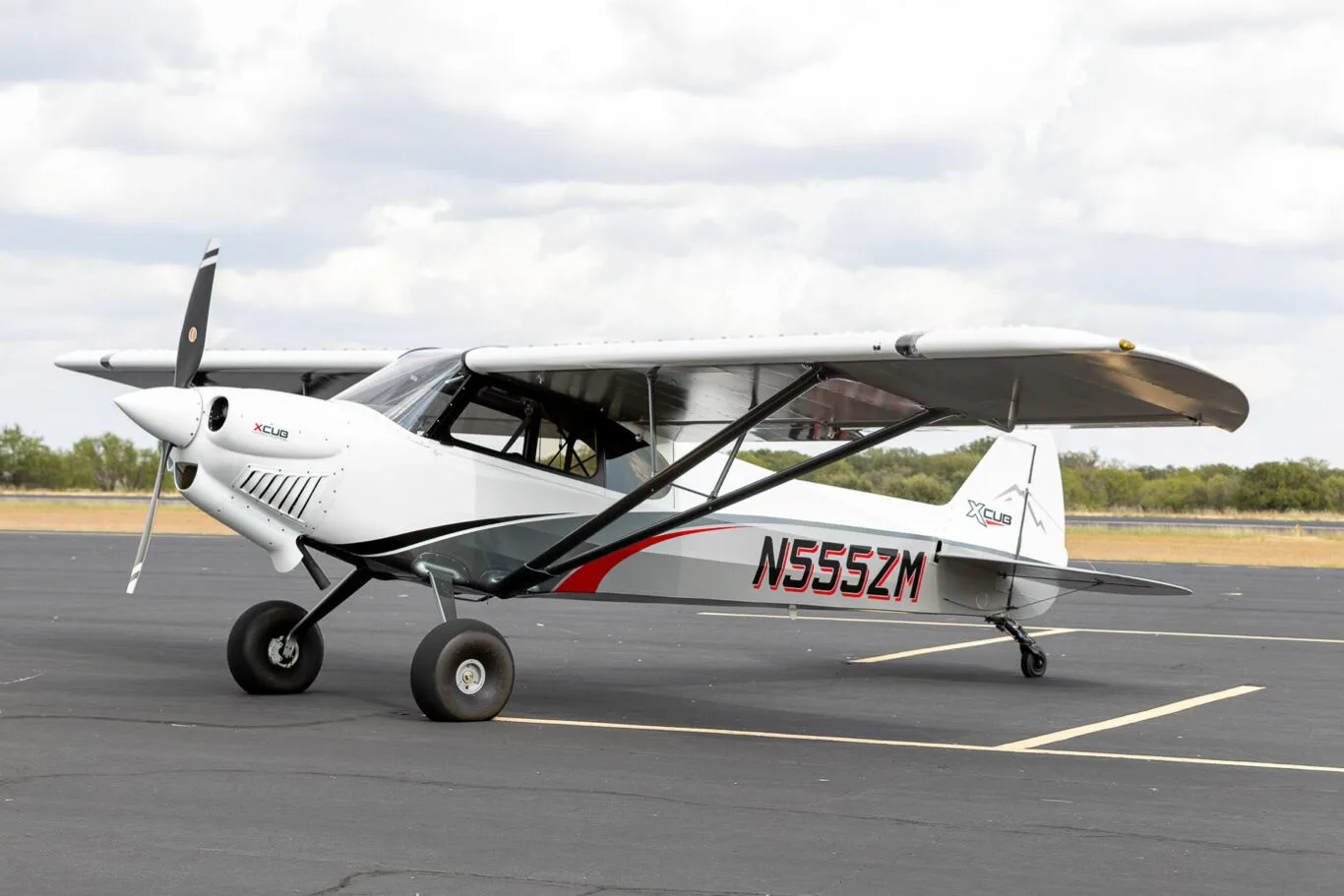 A white and grey X-Cub aircraft with red accents and registration N555ZM parked on an asphalt runway under a cloudy sky.