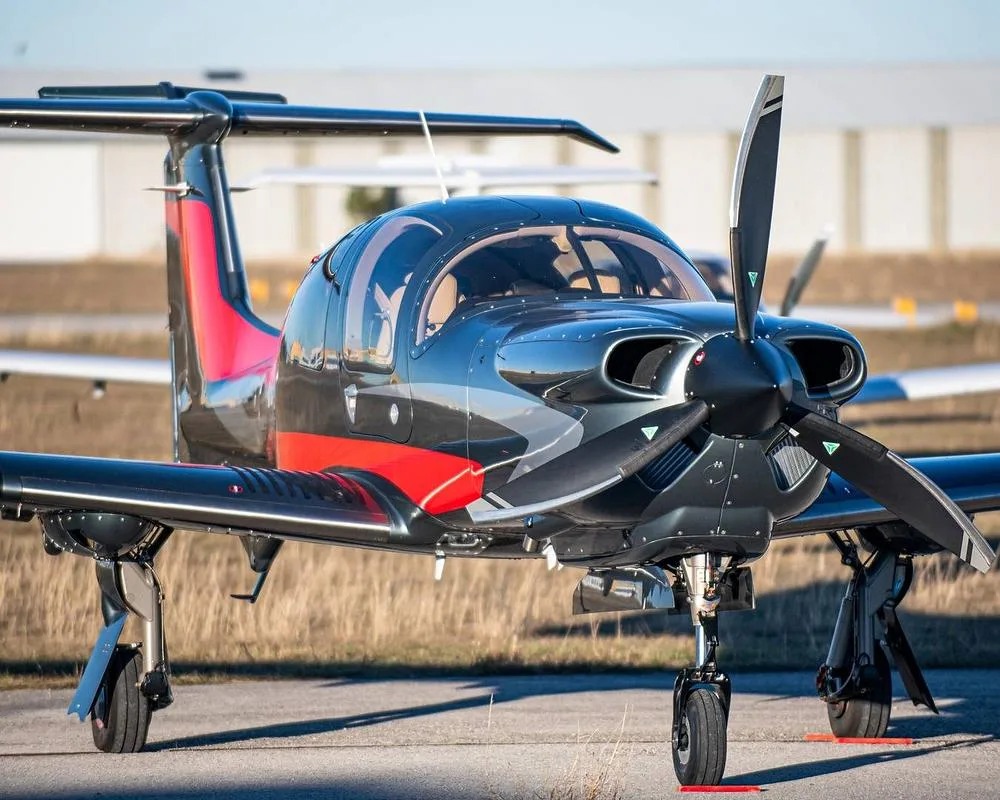 A dark grey and red Diamond DA62 aircraft is parked on an airfield, viewed from the front-left.