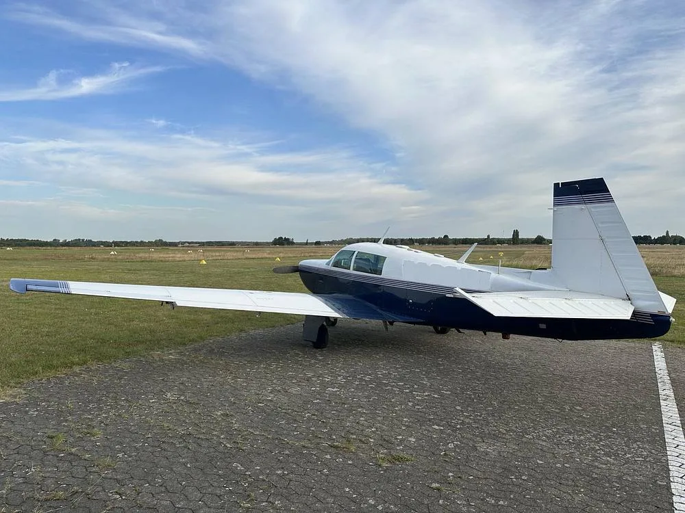 A blue and white single-engine aircraft parked on a tarmac, viewed from the rear-right, with a wide-open field in the background.