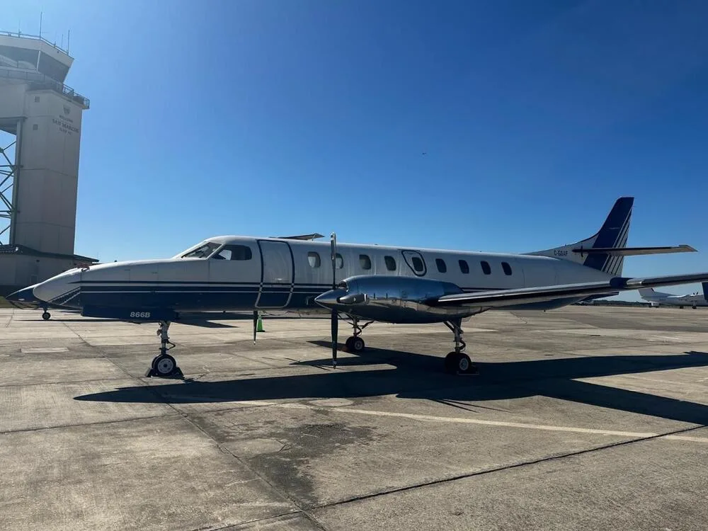 A white and blue Beechcraft 1900 twin-engine turboprop aircraft, registration N-957BA, parked on an airfield next to an air traffic control tower.