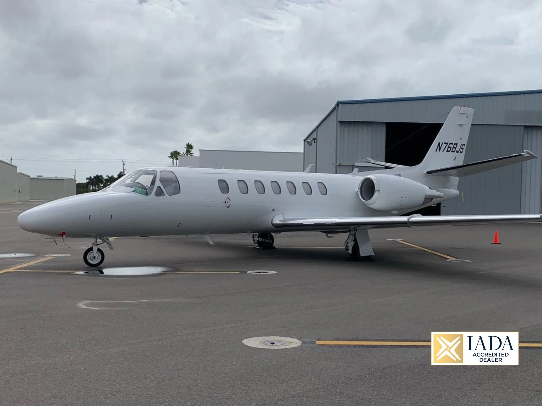 A white Cessna Citation business jet (N768JS) is parked on an airport tarmac next to a hangar under a cloudy sky.