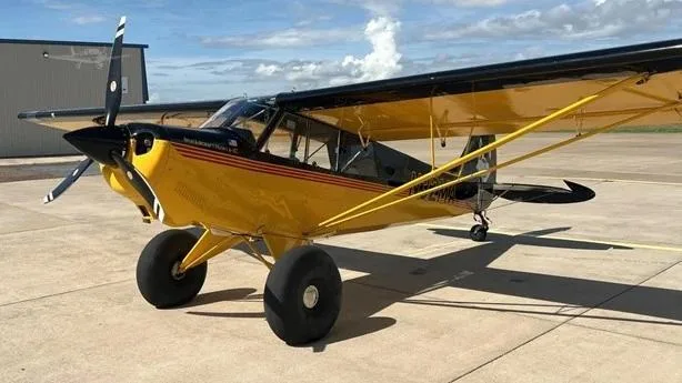 A yellow and black Aviat Husky A-1C aircraft with large tundra tires is parked on a concrete tarmac, viewed from the front-left.