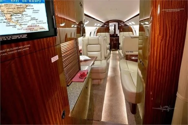 The luxurious interior cabin of a private jet, featuring beige leather seats, dark wood paneling, and a display screen showing a map.