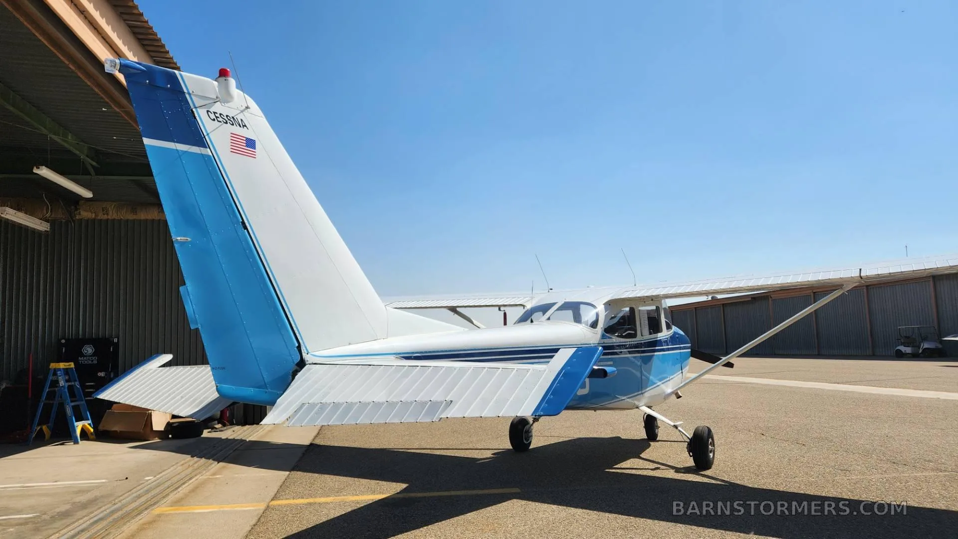 A blue and white Cessna aircraft is parked on an airfield, viewed from the rear-right, with a hangar in the background.