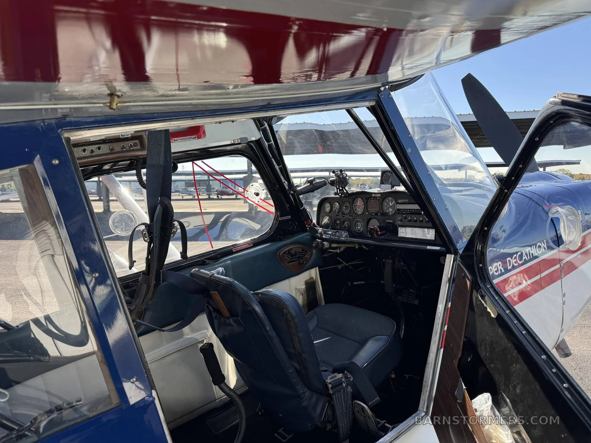 The interior cabin of a Super Decathlon aircraft with a blue seat, showing the open door and part of the cockpit.