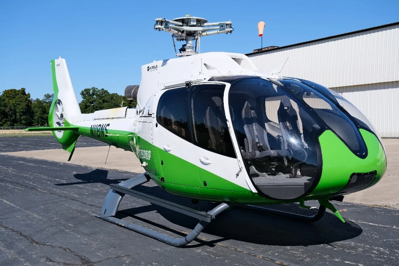 A white and green Airbus H125 helicopter (N111US) is parked on an asphalt tarmac with a building and trees in the background.