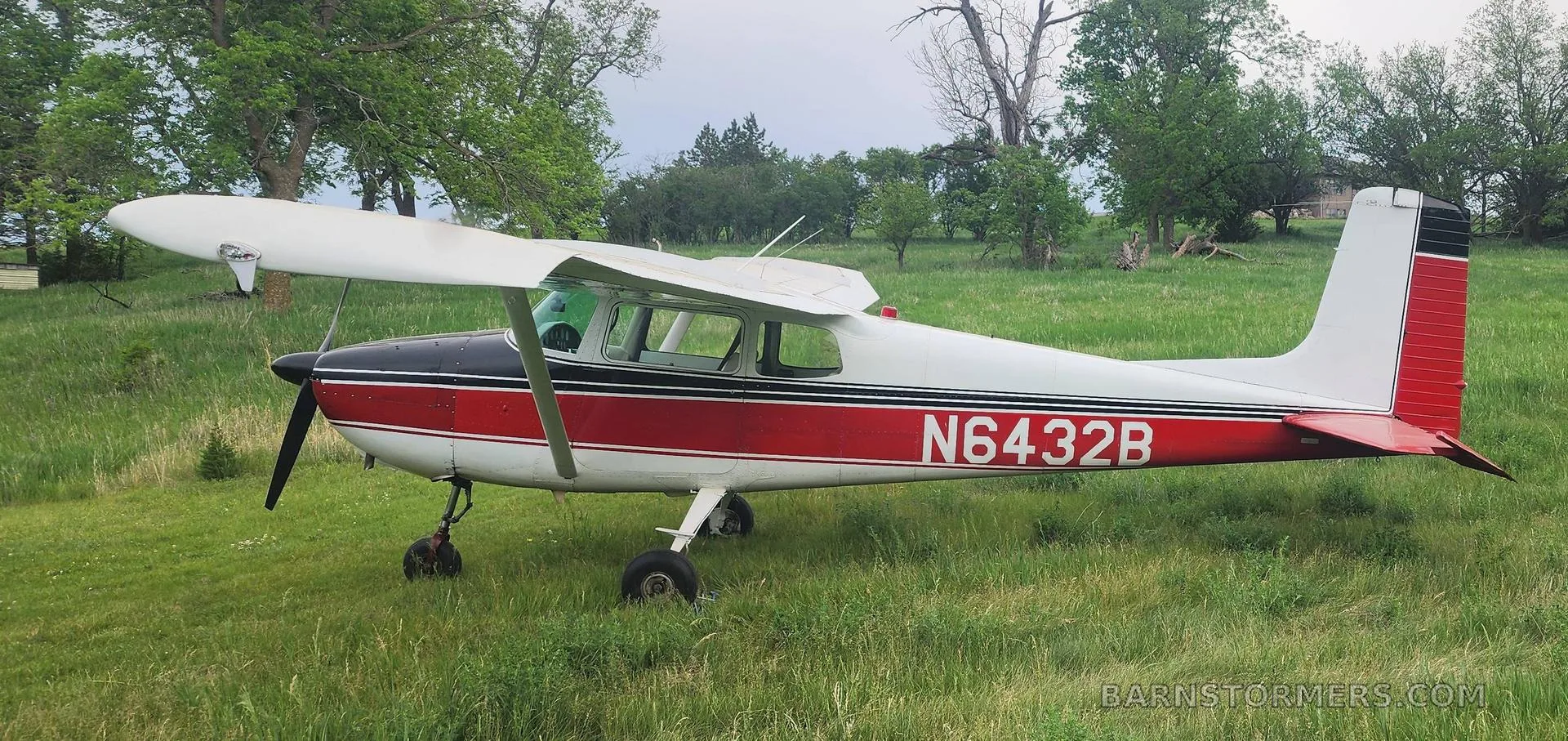 A side view of a white and red Cessna 172 with registration N6432B on a grassy field with trees in the background.