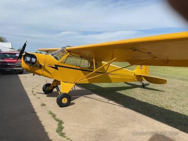 A full side view of a yellow Piper Cub-style aircraft parked on a concrete pad next to a red truck and building.