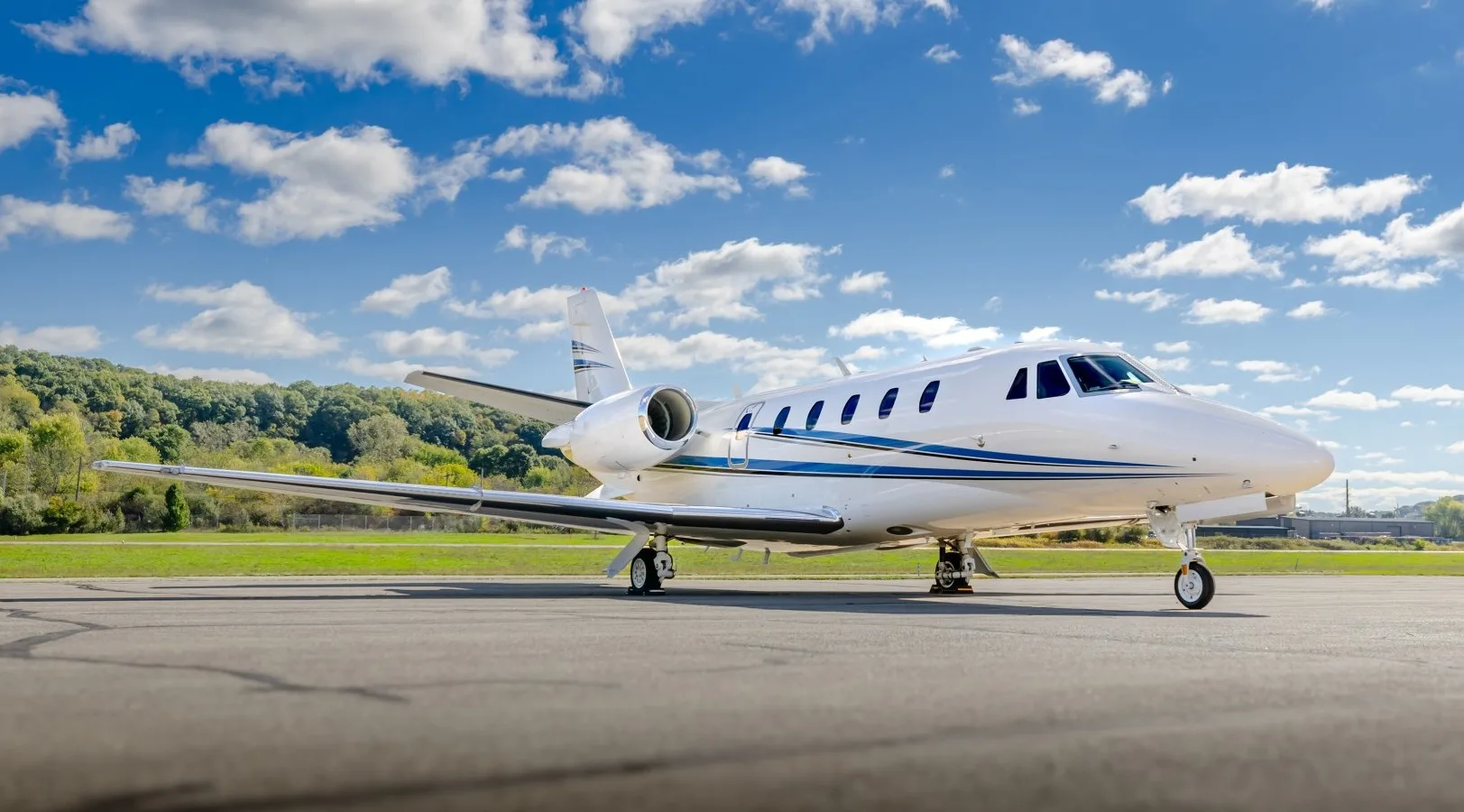 A white Cessna Citation business jet with blue stripes parked on a tarmac under a partly cloudy sky.