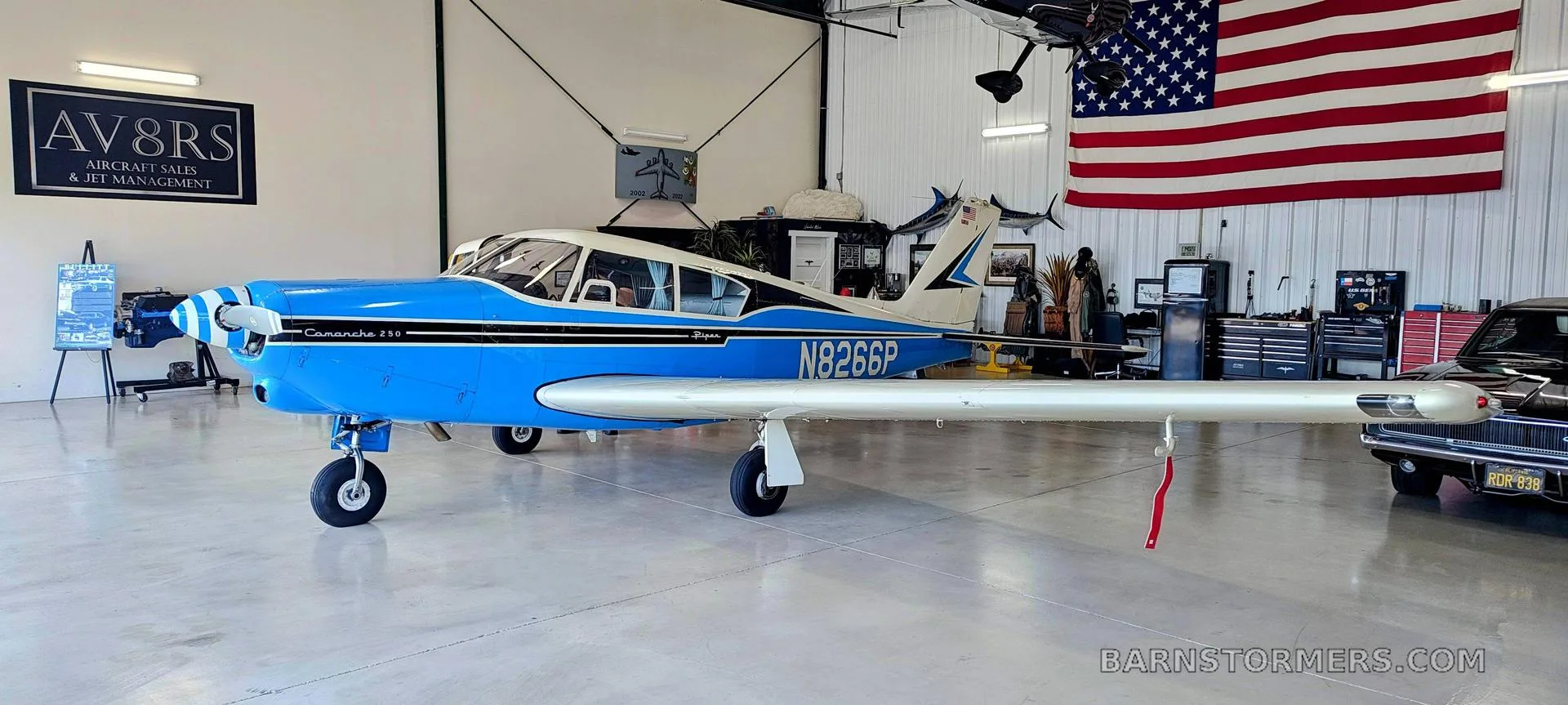 A blue and white Piper Comanche 250 (N8266P) is parked in a hangar with an American flag in the background.