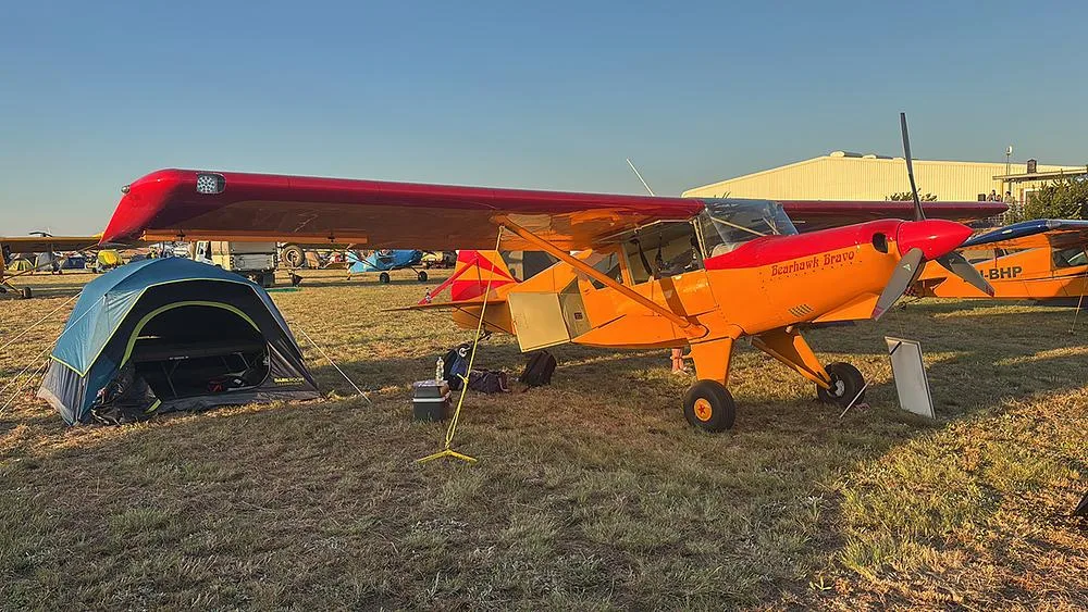 A red and orange Bearhawk Bravo aircraft is parked on a grassy field next to a blue tent at sunset.