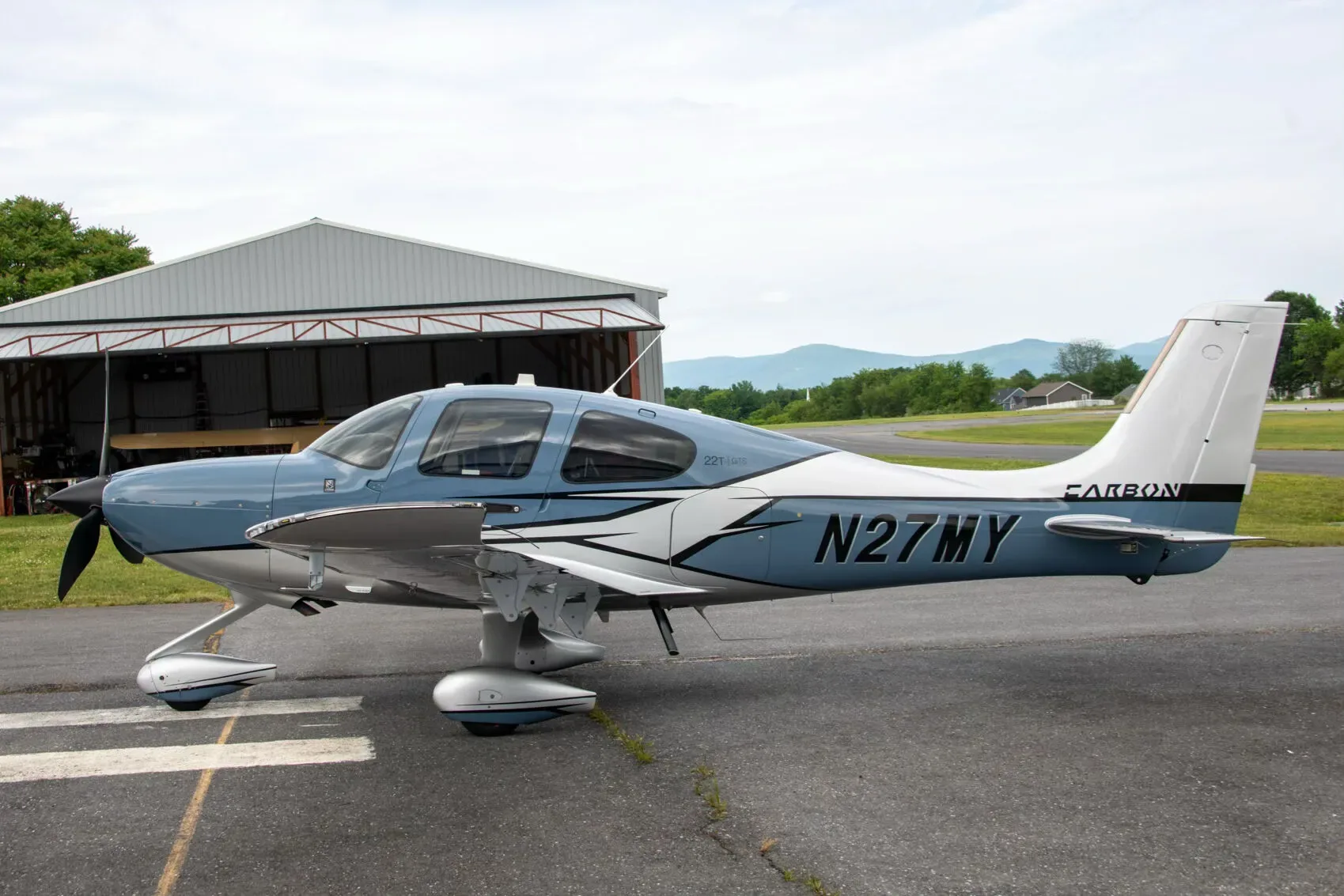 Side view of a blue and white Cirrus SR22T Carbon aircraft, N27MY, parked near an open hangar with mountains in the distance.