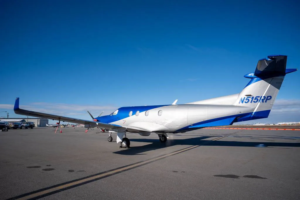 A rear-side view of a blue and silver Pilatus PC-12 aircraft parked on an airfield under a clear blue sky.
