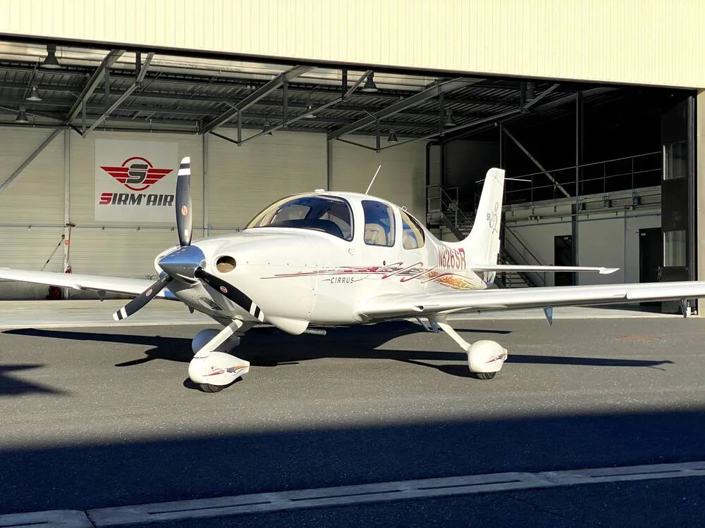 A front-quarter view of a white Cirrus SR22 aircraft with red and gold accent stripes, registration N826SR, parked in front of a hangar.