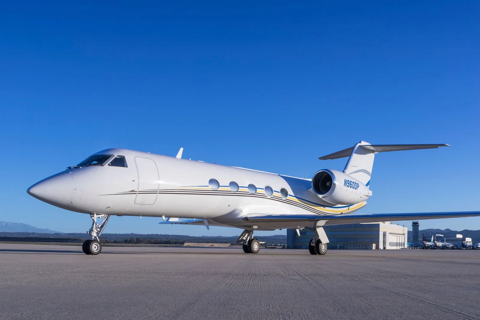 A white Gulfstream business jet with black, yellow, and blue stripes is parked on an airport tarmac under a clear blue sky.