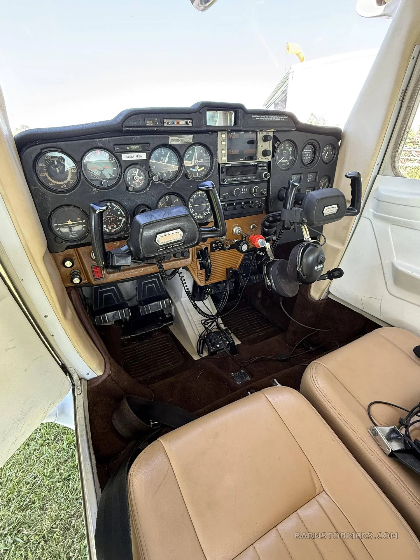 The cockpit of a small aircraft features a black instrument panel, dual yokes, and tan leather seats.