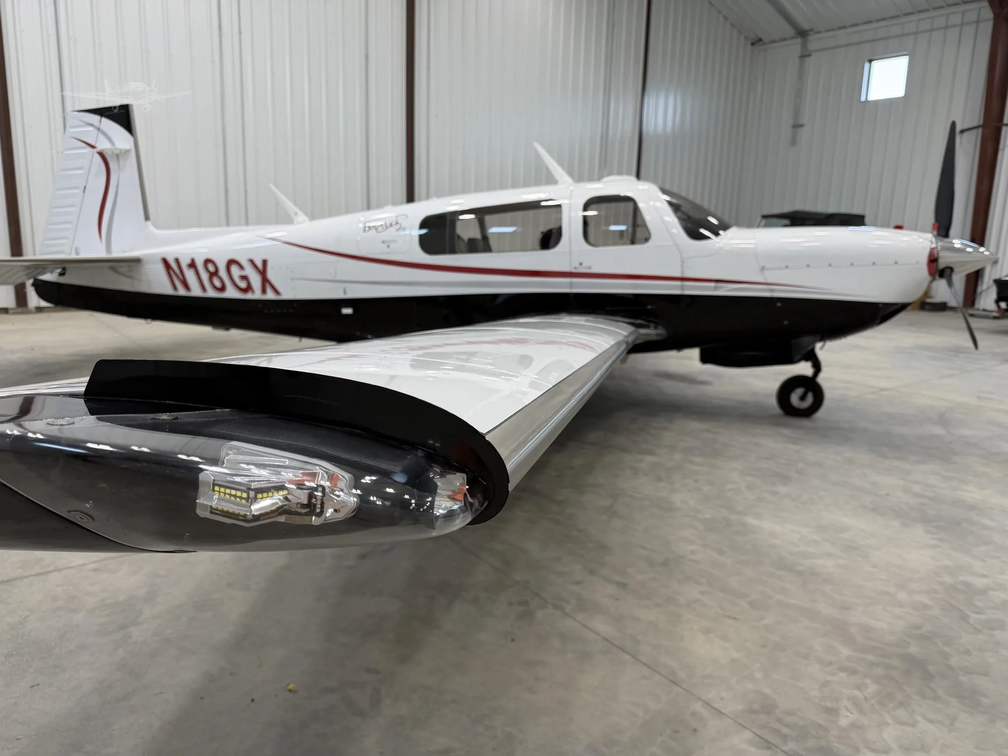 A white and black single-engine propeller aircraft with a red stripe, registration N18GX, is parked in a hangar, viewed from a low angle showing the wingtip light.