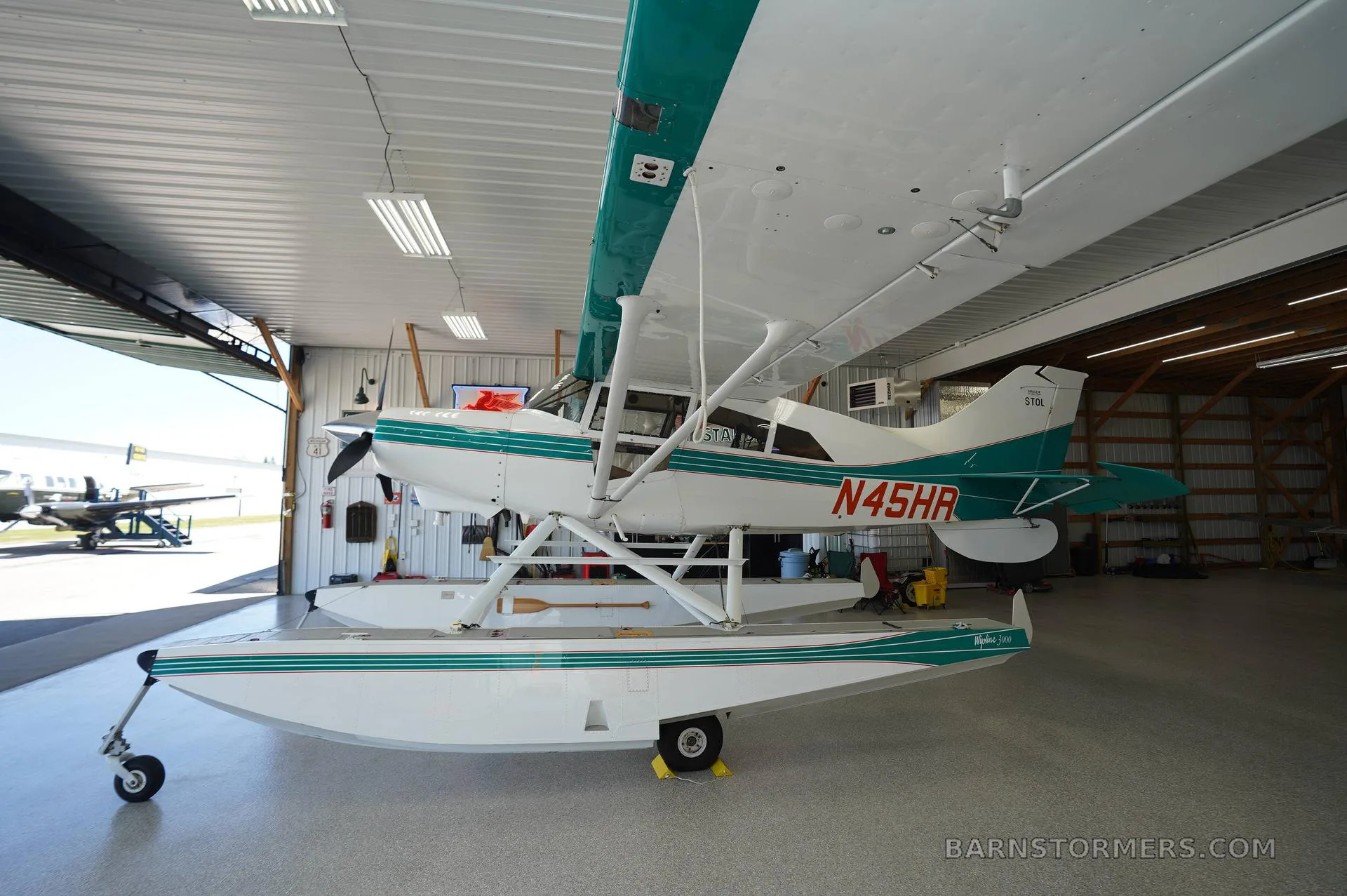 A white and teal floatplane, registration N45HR, is parked inside a hangar, viewed from the front-right.