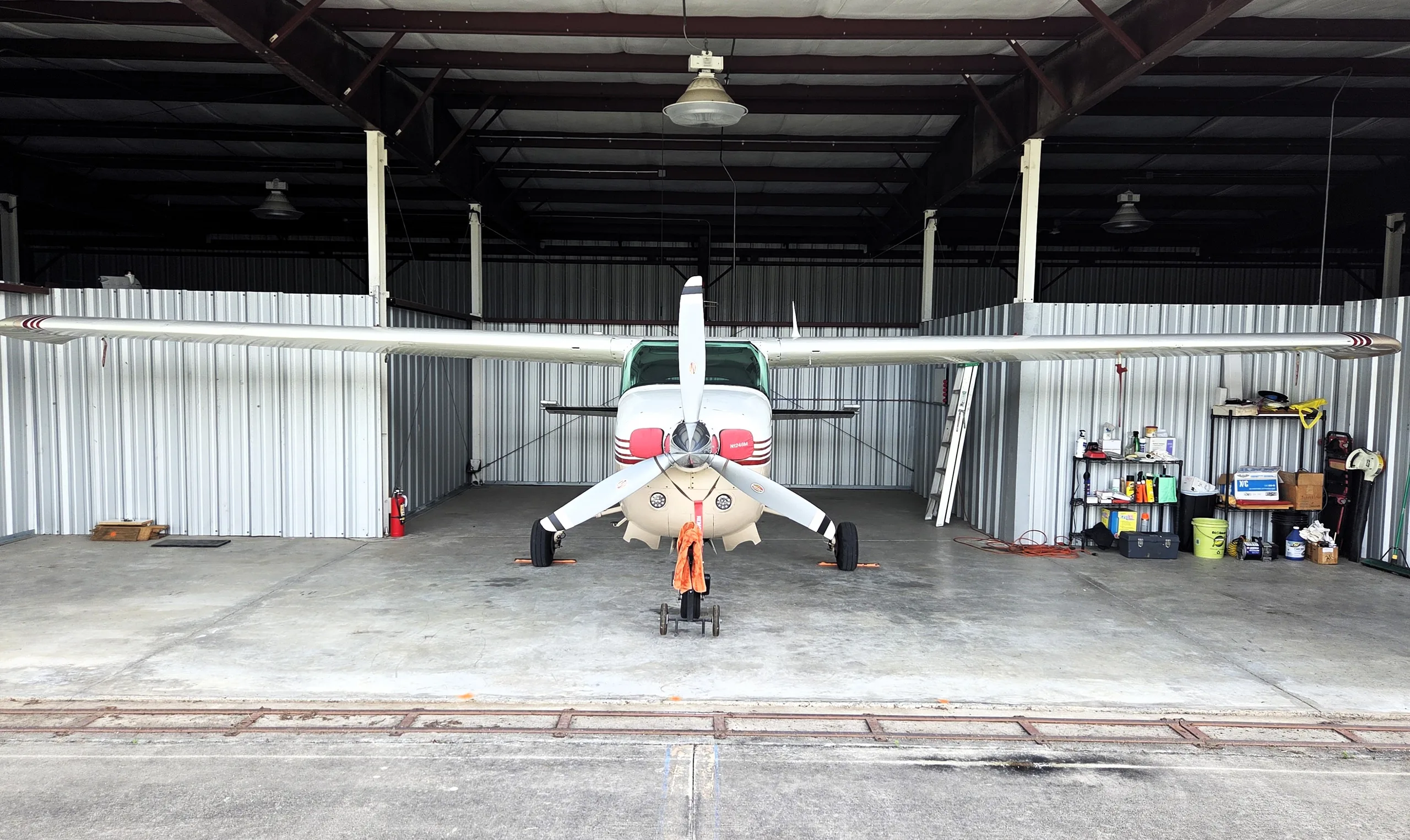 A front view of a white and tan single-engine propeller aircraft with red stripes, parked in a hangar.