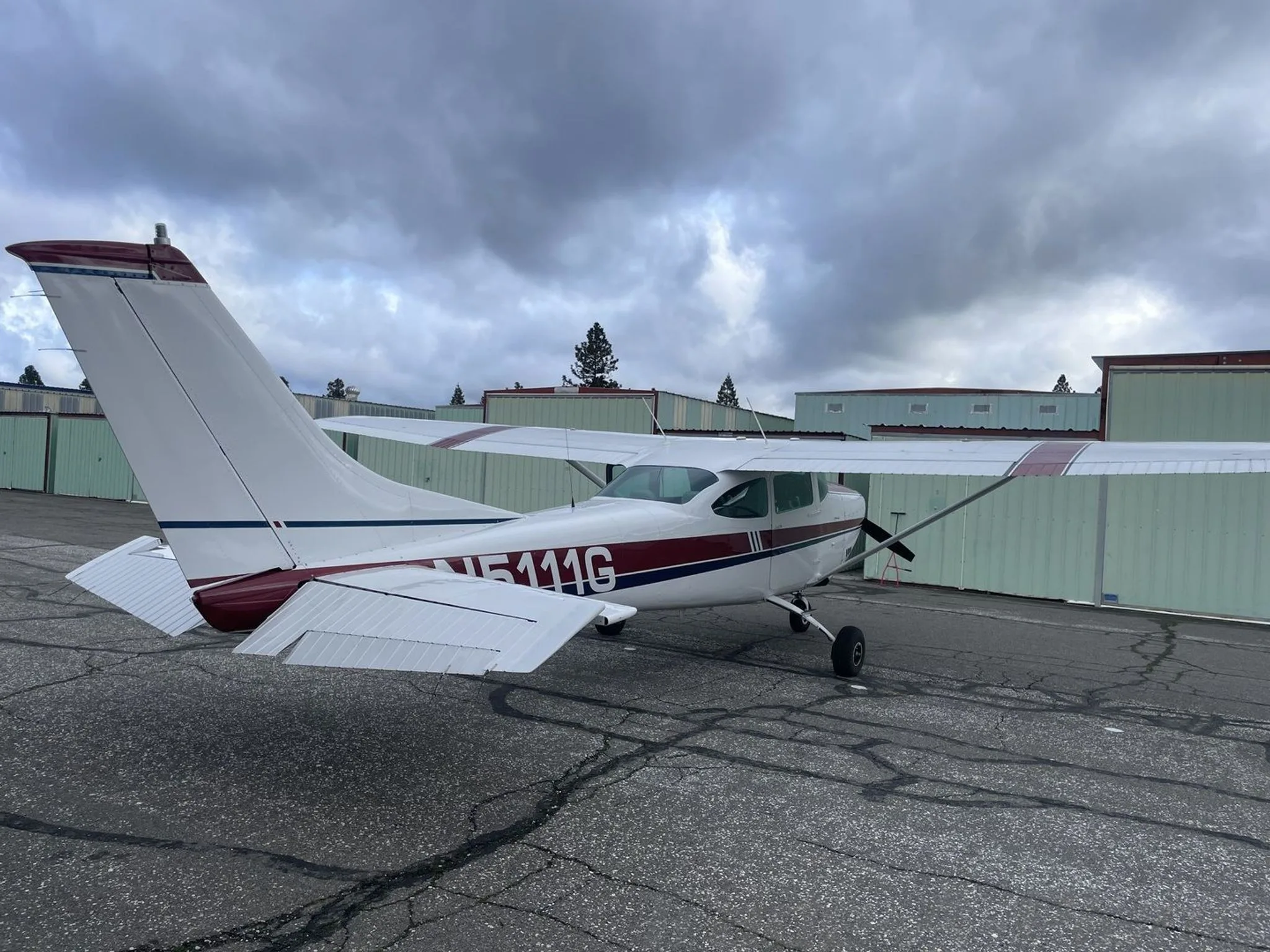 A rear-side view of a white and maroon Cessna 182 aircraft with registration N5111G parked on a tarmac under a cloudy sky.