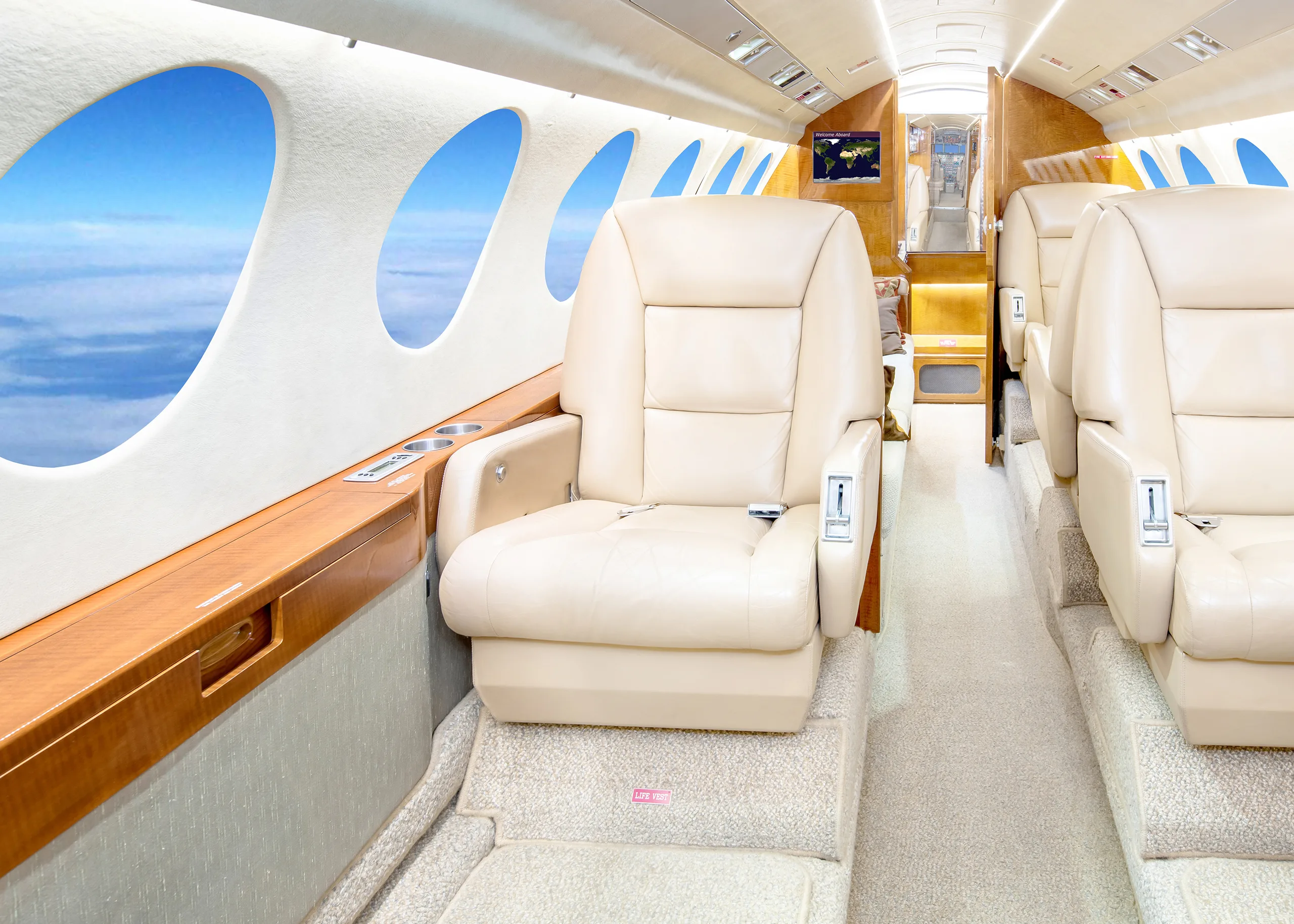A view down the aisle of a private jet cabin, showing a beige leather seat, wood trim, and windows with a sky view.