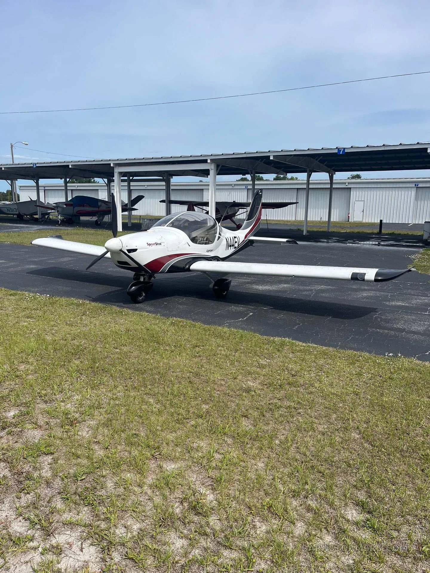 A white and red SportStar aircraft, tail number N44EV, parked under an open-sided hangar structure on a sunny day.