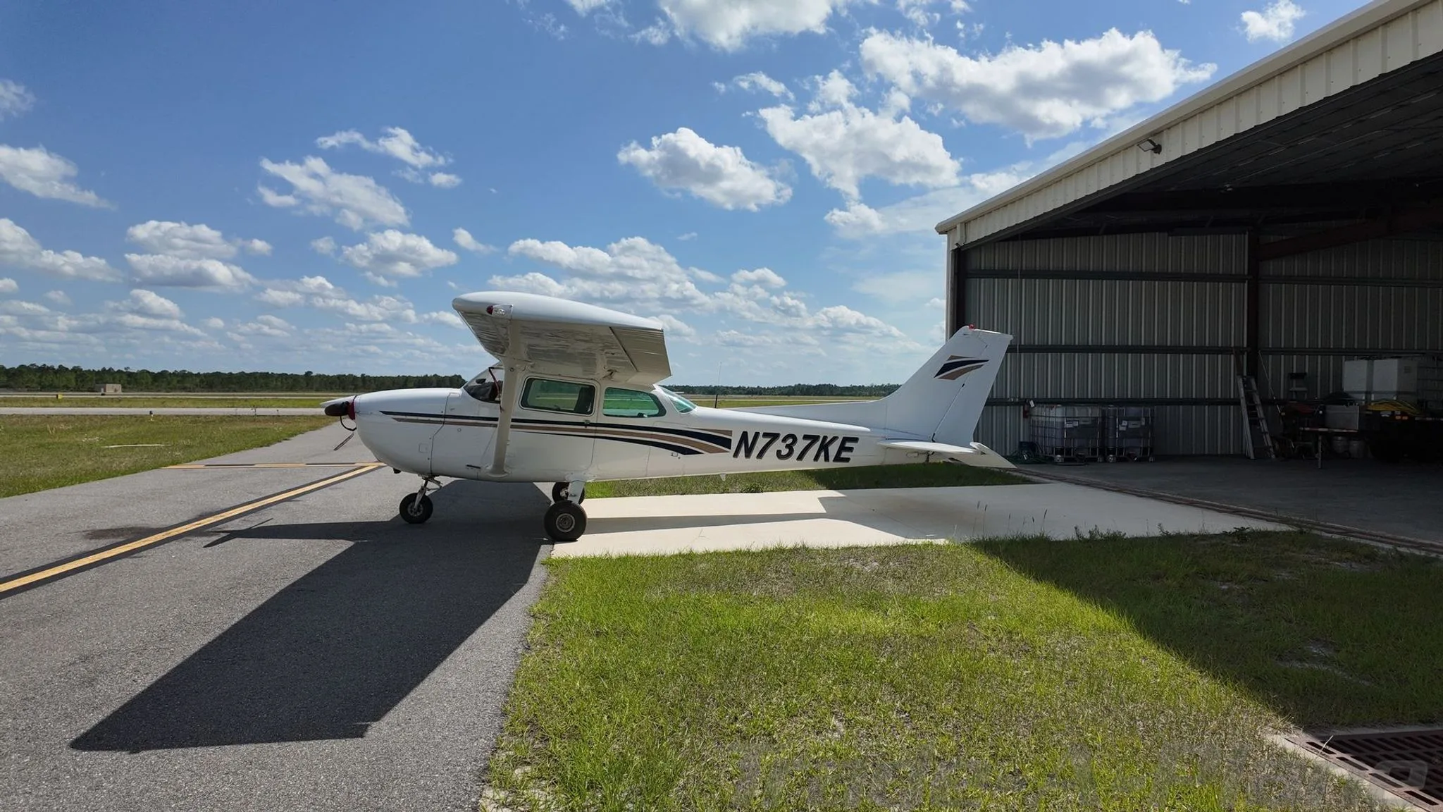 A side view of a white Cessna 172 Skyhawk with brown and blue stripes, tail number N737KE, parked on an airfield next to a hangar.