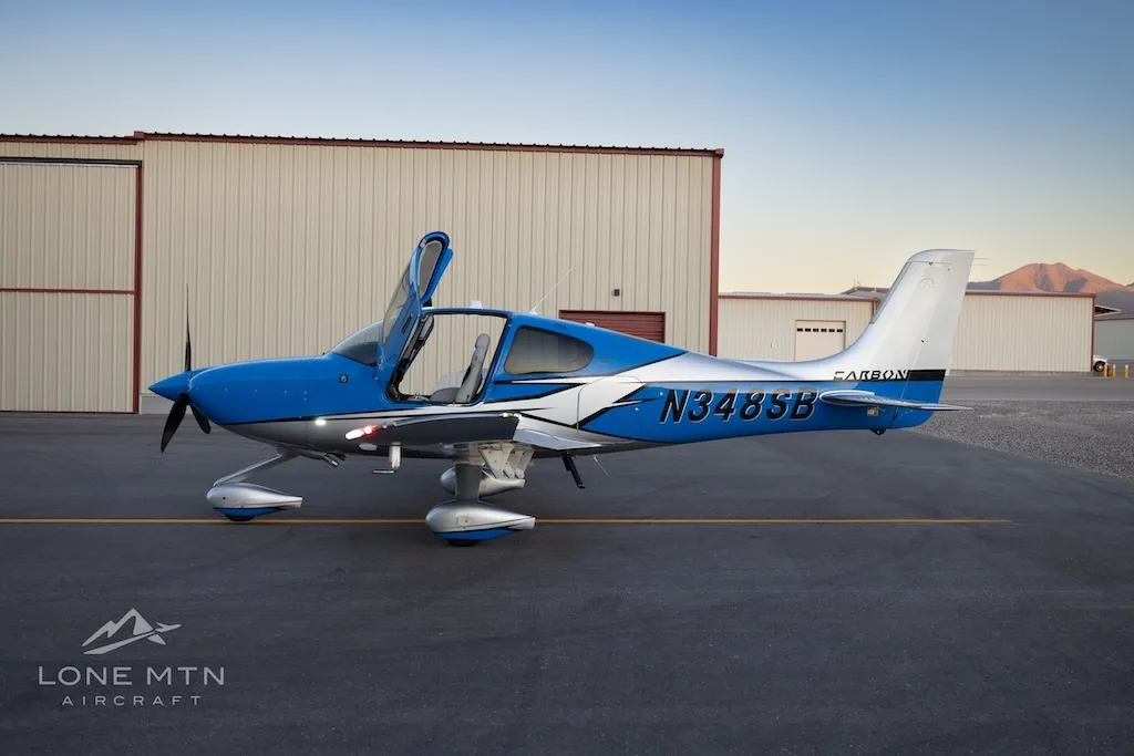 A side view of a blue and silver Cirrus SR22 aircraft with open doors parked on an airfield next to hangars.