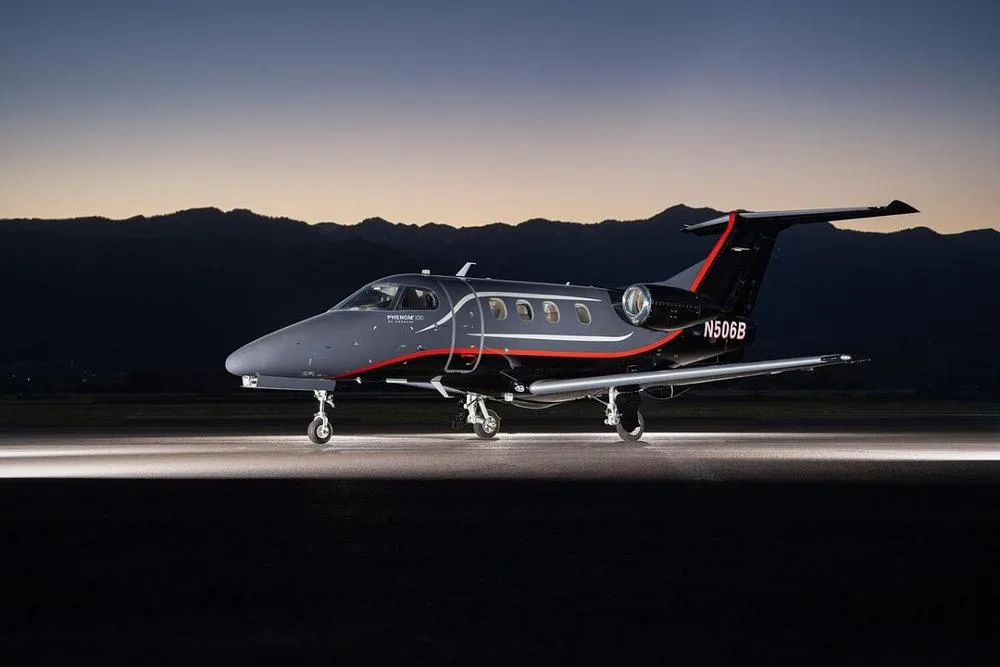 A dark gray and black Embraer Phenom 300 business jet with red accents is parked on a tarmac at dusk, with mountains in the background.