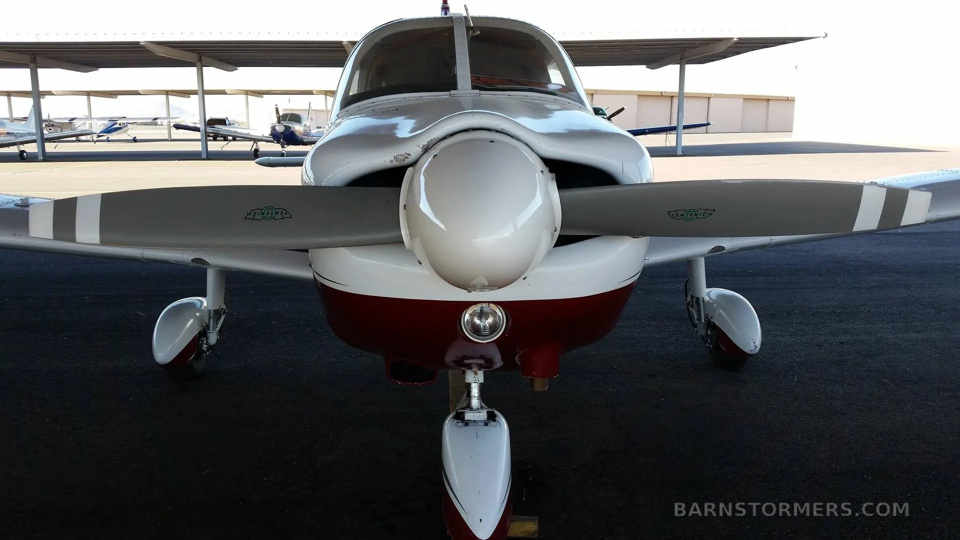 A close-up front view of a white and red Piper Archer II, showing its propeller and nose landing gear under a hangar.