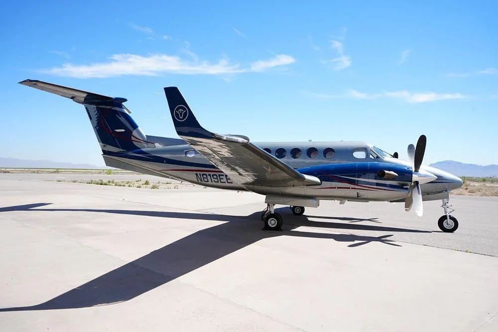A silver and blue twin-turboprop aircraft, N819EE, parked on an airfield with mountains in the background.