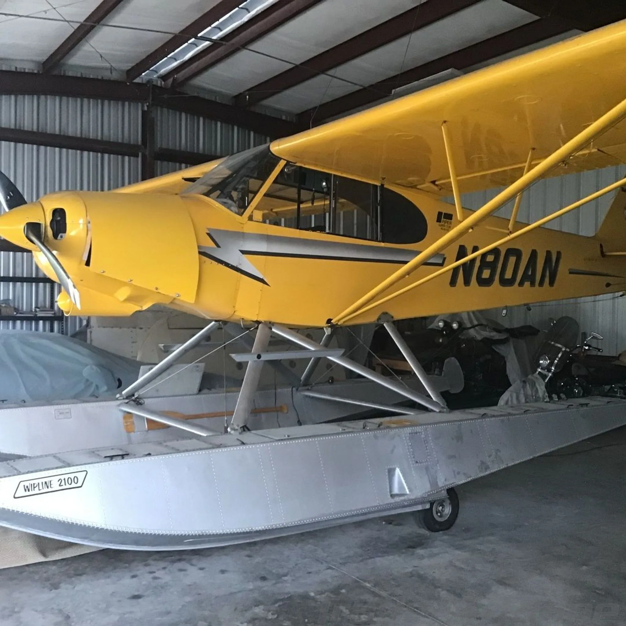 Side view of a yellow Piper Super Cub floatplane (N80AN) with WipLine 2100 floats, parked in a hangar.