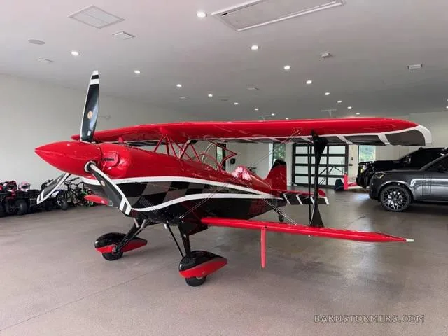 A red and black biplane with a checkered pattern on its fuselage and wings, parked in a garage next to cars and ATVs.