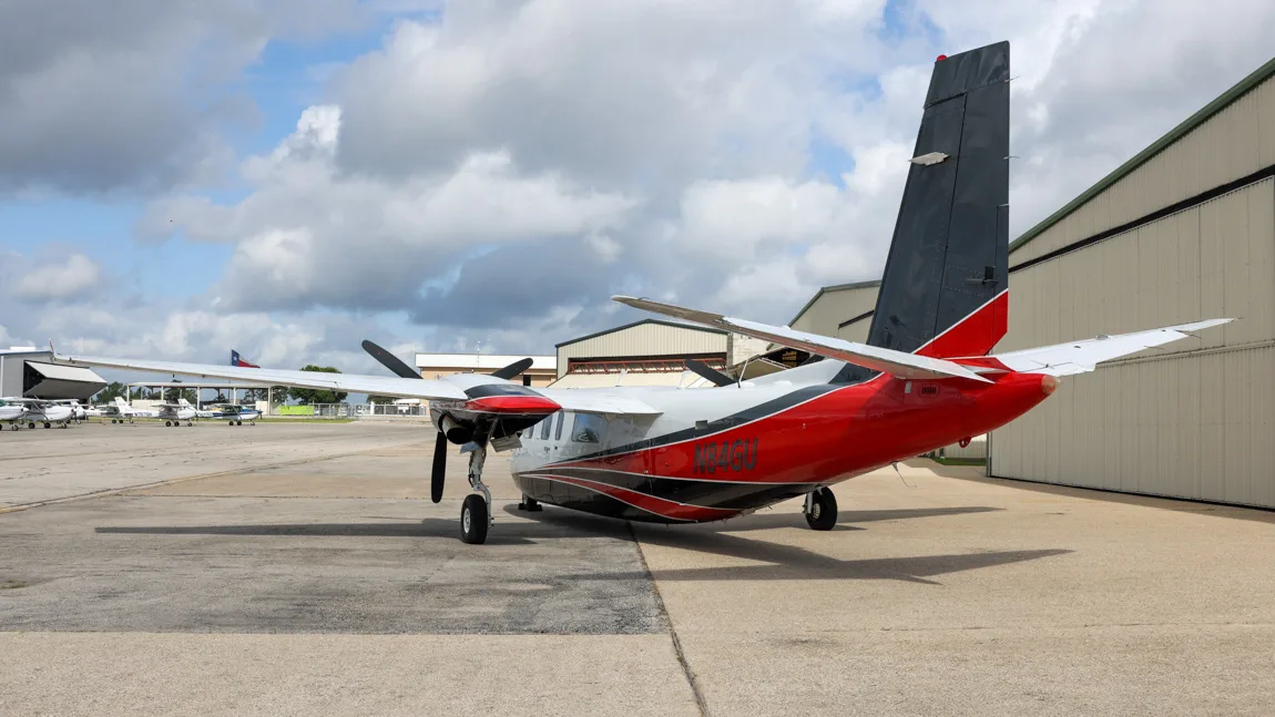 A white, black, and red twin-engine aircraft, N84GU, parked on an tarmac with hangars in the background.