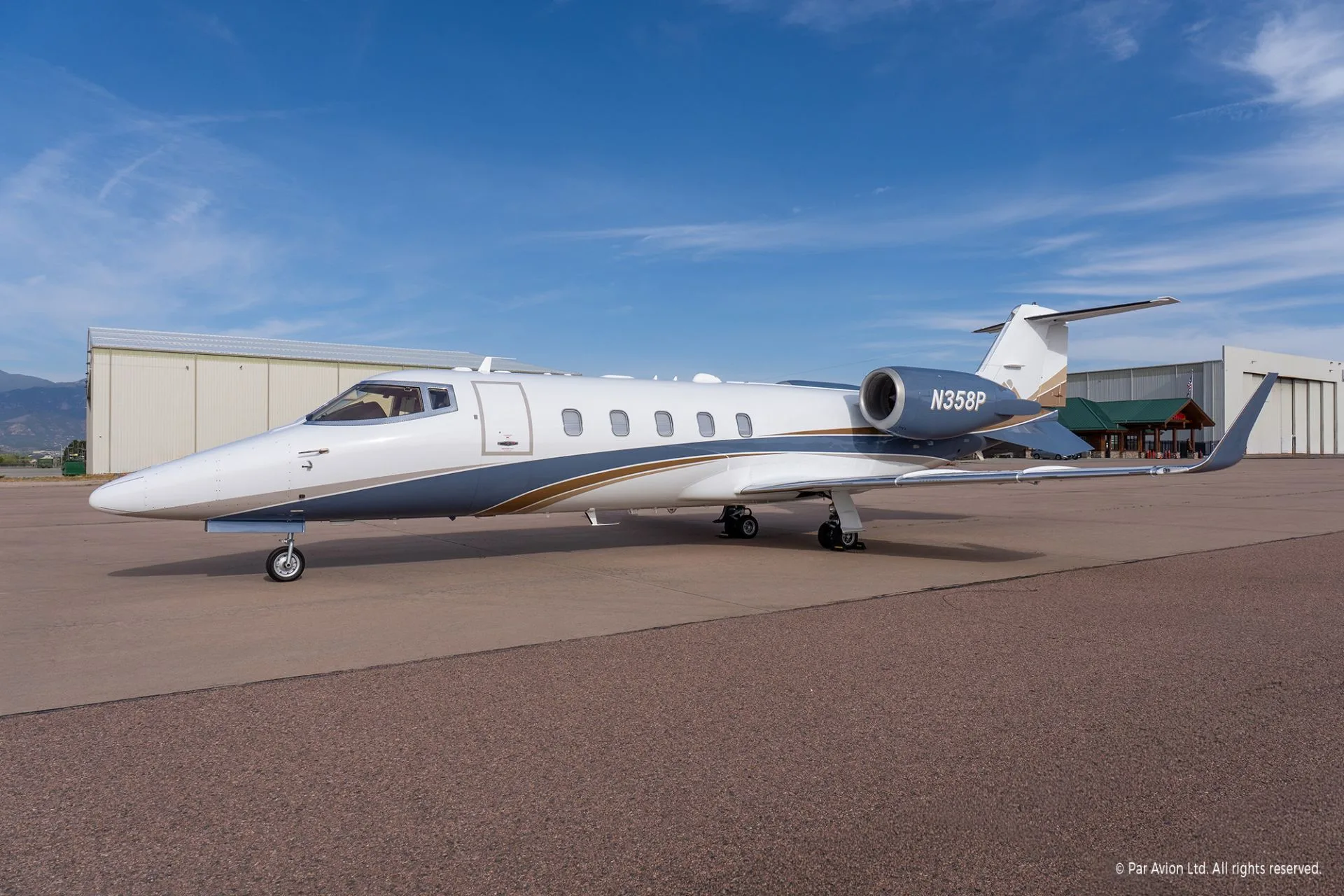 A white, grey, and gold Learjet business jet with registration N358P is parked on an airport tarmac with hangars and mountains in the background.
