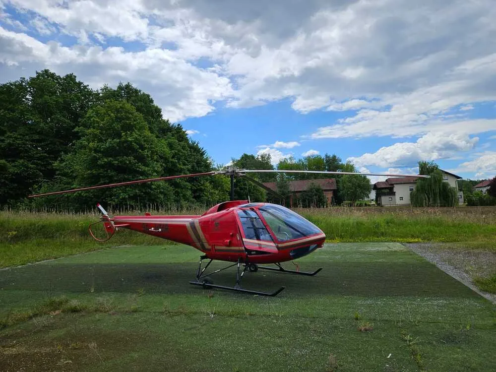 A red helicopter with gold stripes sits on a green helipad under a cloudy sky, with trees and houses in the background.