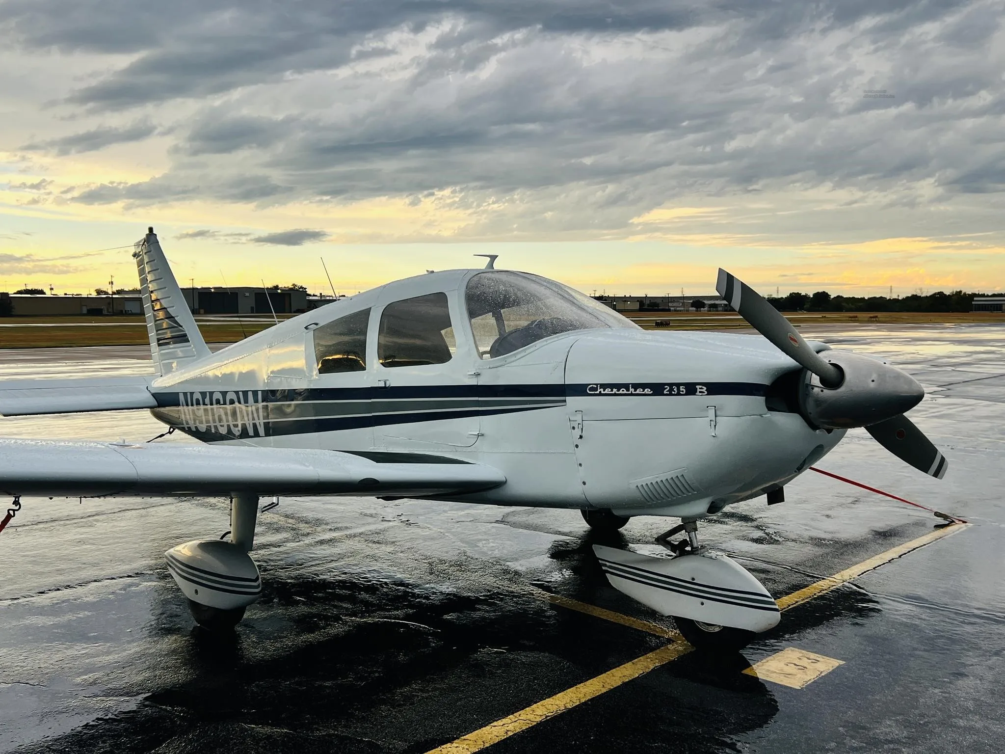 A white Piper Cherokee 235 B aircraft with dark stripes is parked on a wet tarmac under a cloudy sky at sunset.