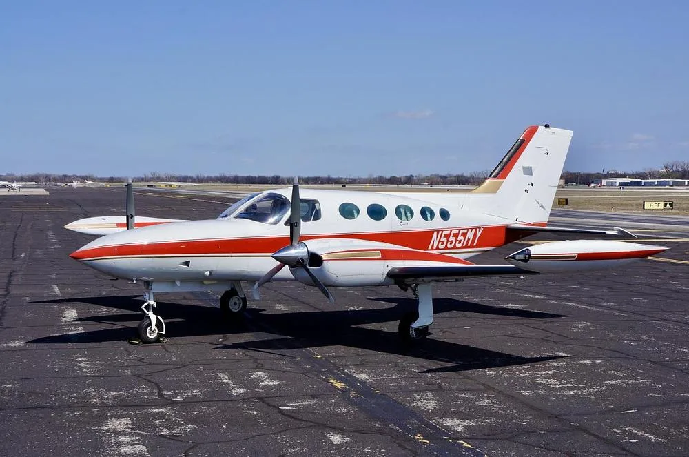A white and red Cessna 400 series twin-engine aircraft, registration N555MY, is parked on an asphalt tarmac under a clear blue sky, viewed from the front-left.