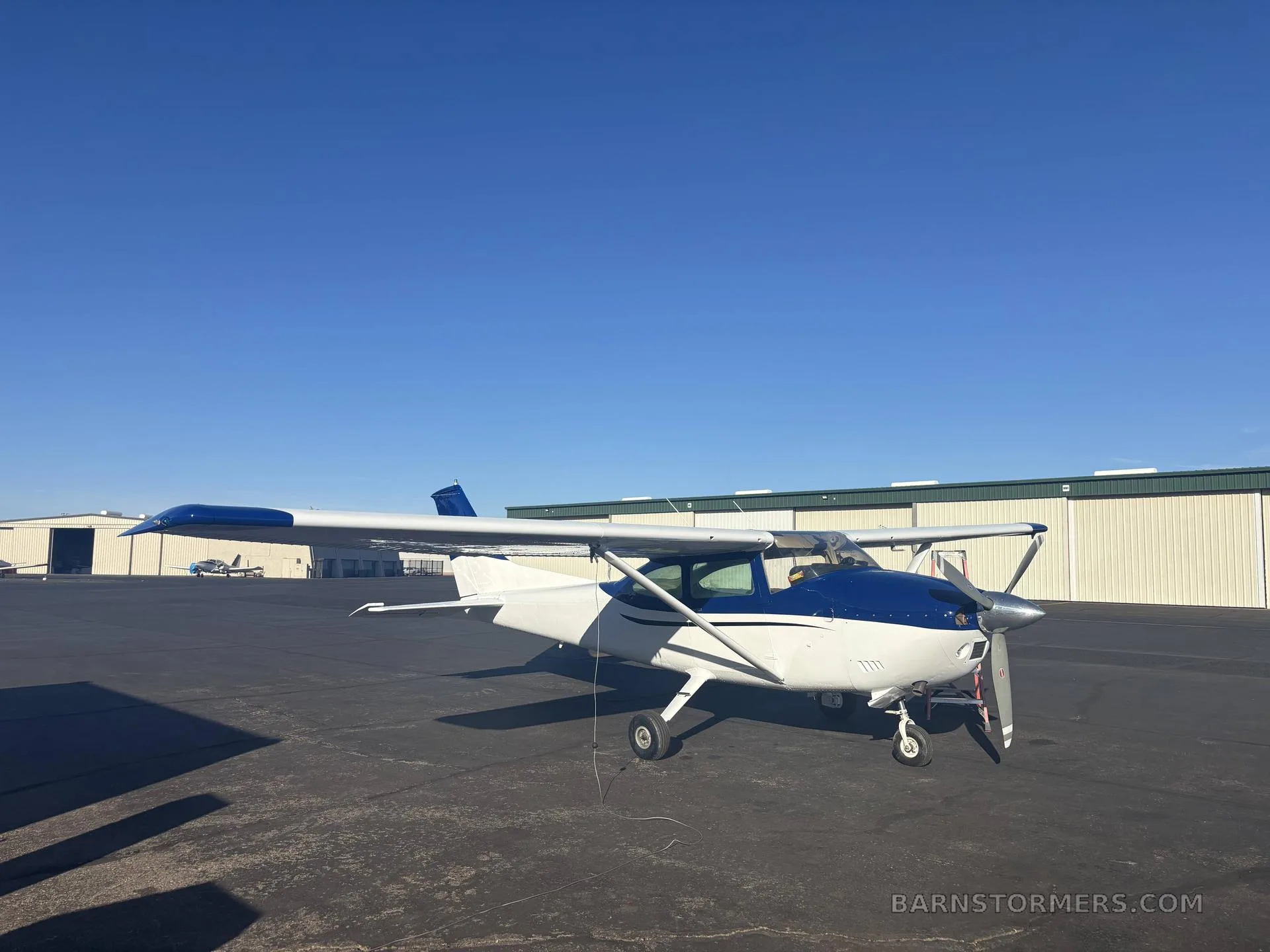 A blue and white single-engine propeller aircraft is parked on an airport tarmac under a clear blue sky.