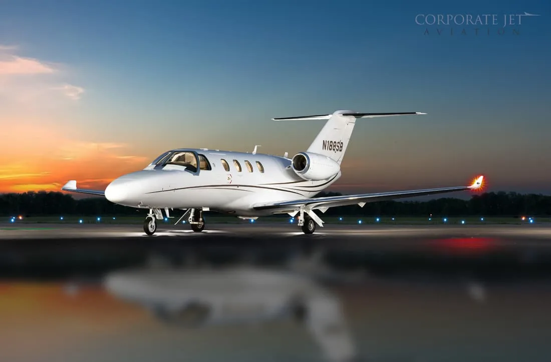 A white business jet (N188SB) with brown stripes sits on a tarmac at sunset, reflecting on the wet ground.
