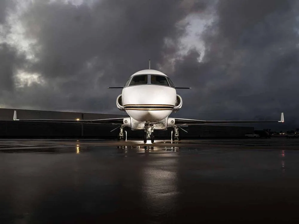A head-on view of a white business jet with gold and black stripes parked on a wet tarmac under a dramatic cloudy sky.