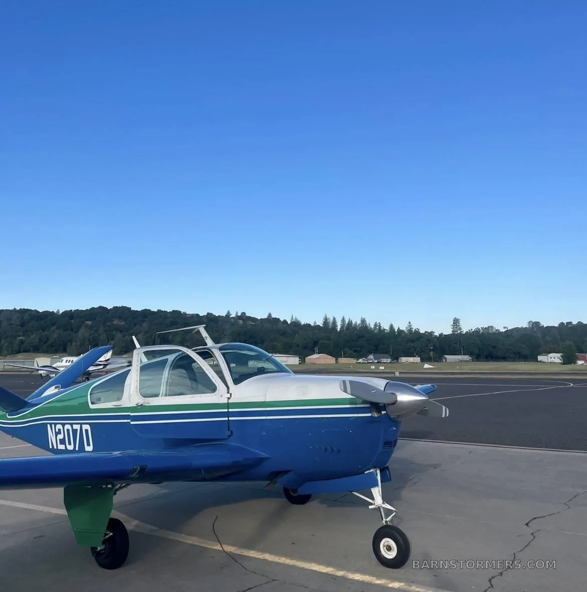 A blue and green single-engine propeller aircraft (N207D) with its canopy open, parked on an asphalt runway under a clear blue sky.