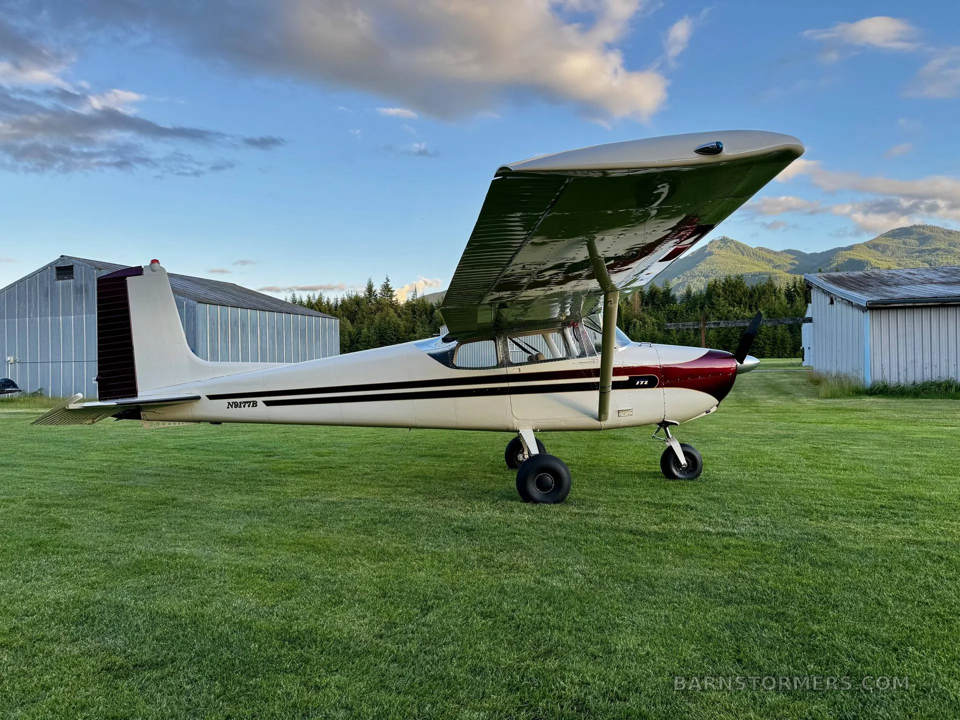 A white and maroon Cessna 172 aircraft is parked on a green grassy field with hangars and mountains in the background under a partly cloudy sky.