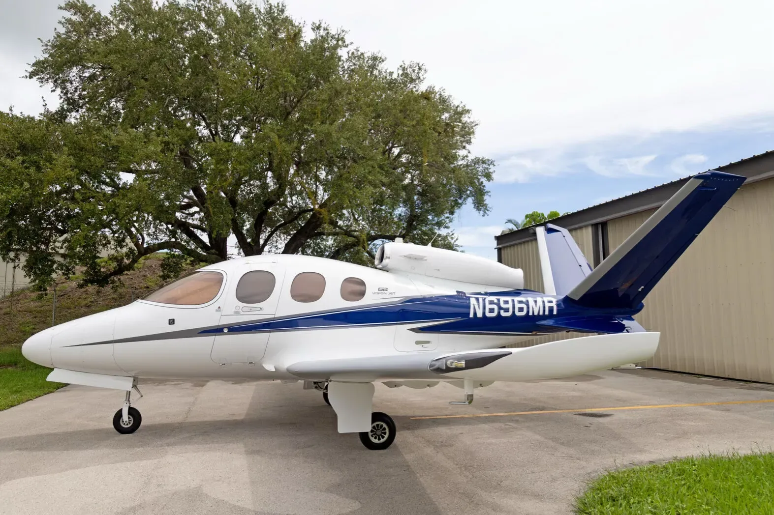 A white Cirrus Vision Jet (N696MR) with blue and grey stripes parked on a tarmac next to a large tree and hangar.