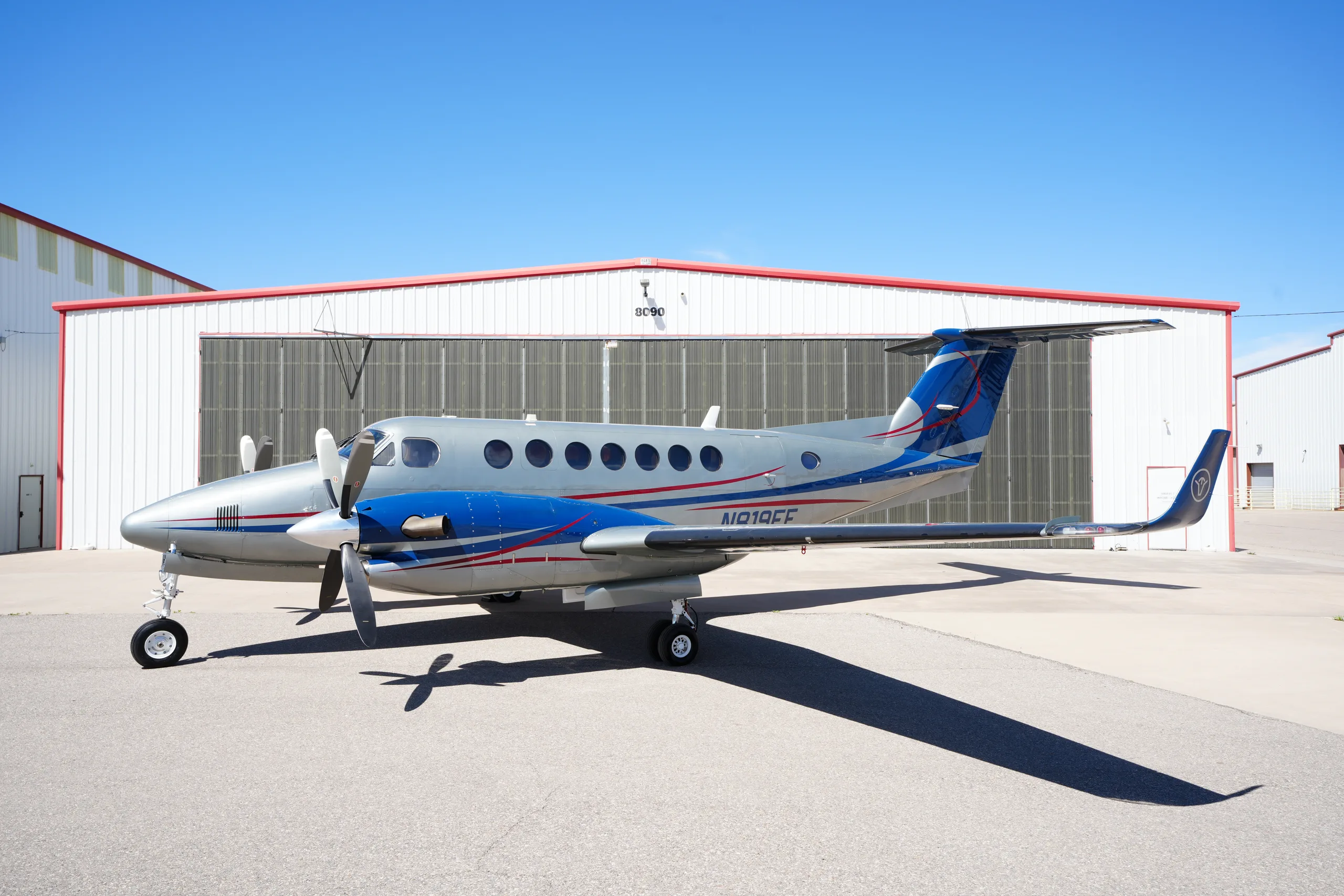 A silver and blue twin-engine turboprop aircraft (N19FF) parked on an airfield in front of a hangar.