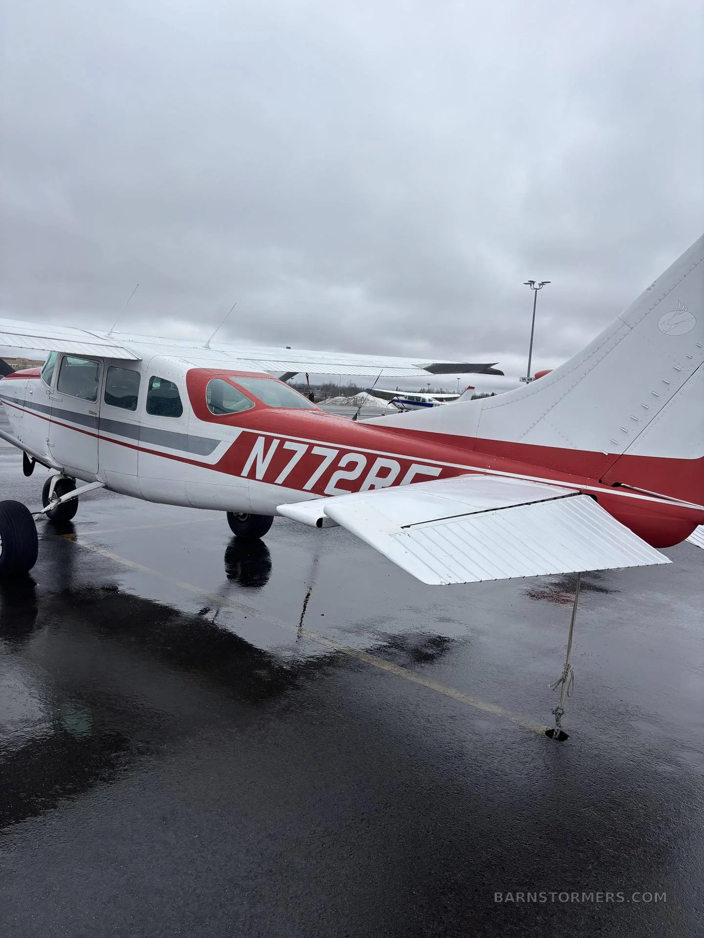 The rear-side view of a white and red single-engine Cessna aircraft, registration N772RF, parked on a wet tarmac.