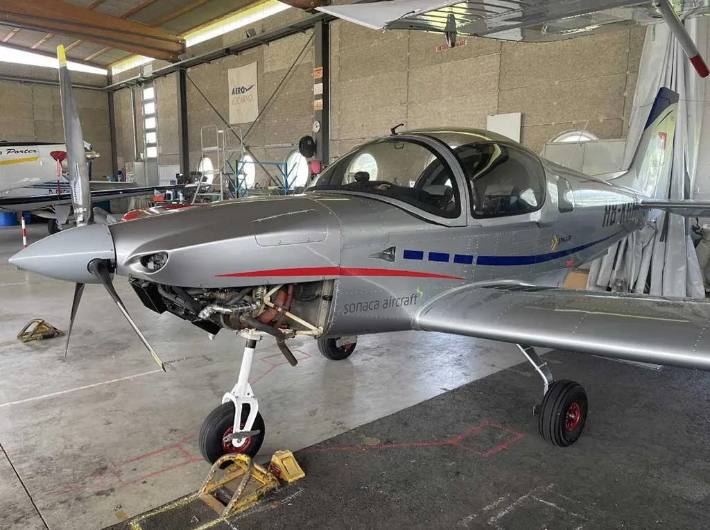 Three-quarter front view of a silver Sonaca 200 aircraft with blue stripes and open engine cowling, parked in a hangar.