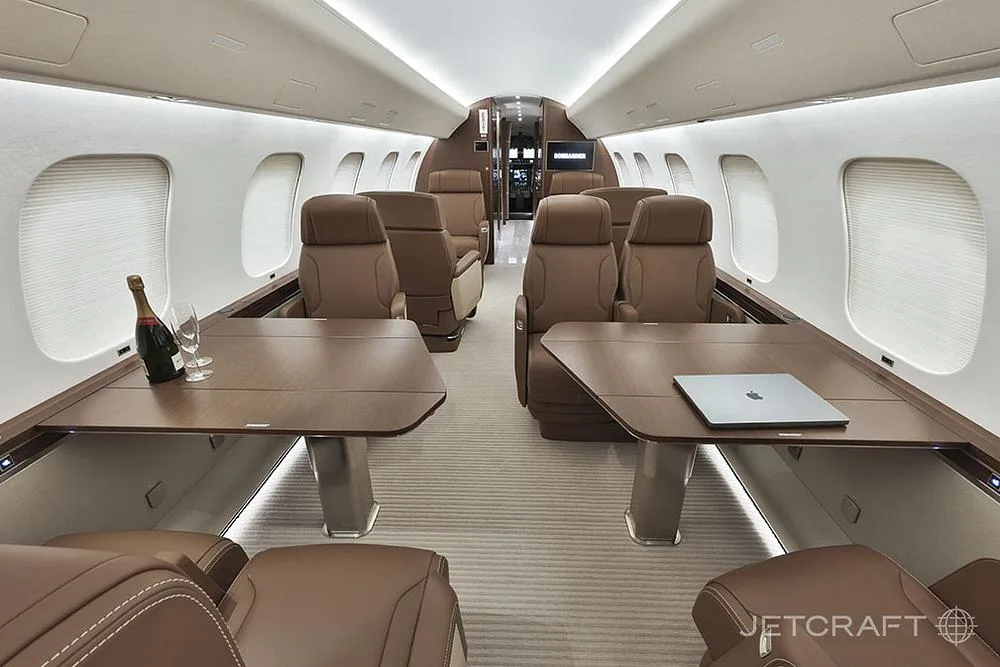 The luxurious interior of a business jet cabin features brown leather seats, two tables, and a bottle of champagne.